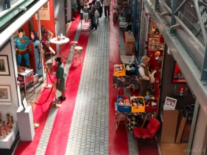 Cyrille · 25/04/2026 · 5148 x 3860 Cette photographie montre une allée commerçante animée. Le sujet principal est une rue piétonne avec des boutiques et des passants.La composition est symétrique, encadrée par des magasins de chaque côté et une large bande rouge au centre. Le cadrage est large, capturant une vue plongeante depuis un étage supérieur.La lumière est naturelle et diffuse, éclairant uniformément l’espace. Les couleurs dominantes sont les tons neutres (beige, gris) avec des touches vives (rouge, jaune). L’ambiance est dynamique et urbaine, typique d’un centre commercial ou d’une galerie marchande.
