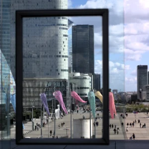Fabienne · 3670 x 3670 Cette photographie montre une vue urbaine prise à travers une fenêtre ou un cadre noir. Le sujet principal est un ensemble de buildings modernes reflétant la lumière, avec des structures géométriques et des façades en verre.La composition met en avant un espace public en contrebas, où des parasols colorés (rose, violet, bleu) sont disposés. Le cadrage est serré, centré sur ces éléments. La lumière est naturelle, claire et légèrement contrastée, accentuant les teintes vives. Les couleurs dominantes sont les gris des bâtiments, les tons pastel des parasols et le bleu du ciel.L’ambiance est dynamique et contemporaine, mêlant modernité architecturale et vie urbaine.
