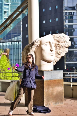 Fabienne · 28/03/2026 · 3016 x 4524 Cette photographie met en scène une femme comme sujet principal, posant devant une tête sculptée antique. La composition joue sur le contraste entre le moderne (bâtiments en arrière-plan) et l'antique (la sculpture). Le cadrage est serré sur la femme et la tête, avec une profondeur de champ qui met en valeur les détails.La lumière est naturelle et semble provenir d’un angle latéral, créant des ombres douces. Les couleurs dominantes sont neutres (beige, gris) avec des touches de vert et bleu. Le style est documentaire et spontané, évoquant une ambiance à la fois urbaine et culturelle, mêlant histoire et modernité.