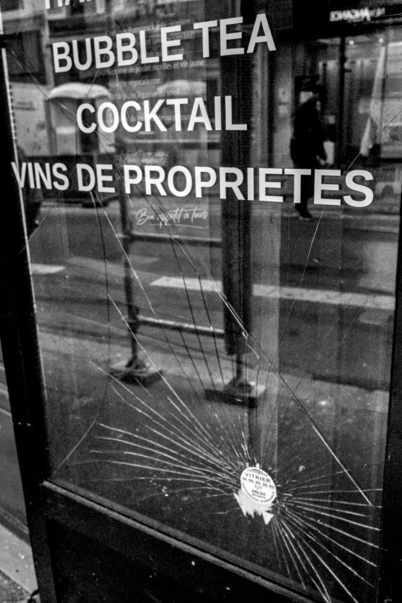 Frédéric · 11/04/2026 · 1121 x 1680 Cette photographie montre une vitrine endommagée avec des éclats de verre rayonnants autour d’un trou central. Le sujet principal est l’inscription partiellement lisible : "Bubble Tea Cocktail Vins de Propriétés".La composition est centrée sur la vitrine, avec un cadrage serré. La lumière, contrastée, accentue les ombres et les reflets, créant un effet dramatique. En noir et blanc, les tons dominants varient du blanc éclatant aux noirs profonds.Le style est documentaire et brut, évoquant une ambiance de désordre et de négligence, voire de vandalisme, renforçant une impression de mélancolie et de rupture.