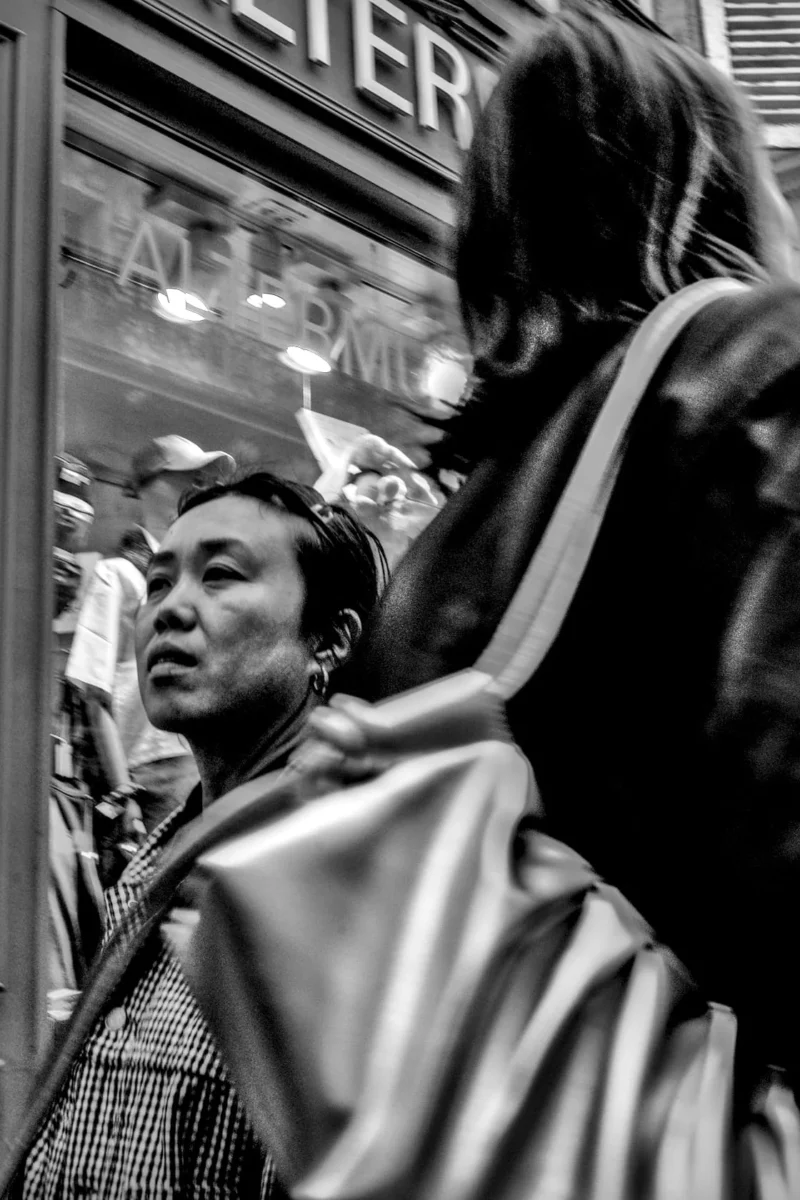 Frédéric · 11/04/2026 · 1120 x 1680 Cette photographie en noir et blanc met en scène deux personnes dont une seule est nette, regardant à travers une vitrine. Le sujet principal semble être la personne au premier plan, avec une expression neutre ou légèrement surprise.La composition joue sur la profondeur de champ, isolant partiellement le visage. Le cadrage est serré, centré sur les visages et la vitrine reflétant des éléments urbains.La lumière est douce, naturelle, créant des contrastes modérés. Les tons dominants sont les gris et noirs, typiques du noir et blanc, renforçant une ambiance intemporelle et urbaine. Le style est documentaire et spontané, évoquant une scène de vie quotidienne.