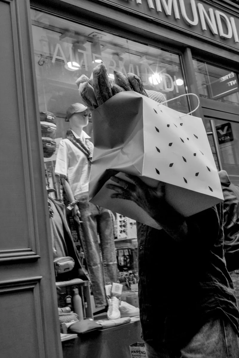 Frédéric · 11/04/2026 · 1120 x 1680 Cette photographie en noir et blanc met en scène une personne tenant un sac blanc à motifs géométriques. Le sujet principal semble être cette silhouette floue, partiellement cachée par le sac.La composition utilise un cadrage serré sur la vitrine d’un magasin, avec des éléments urbains en arrière-plan. La lumière est douce, avec des reflets et des ombres contrastées, créant une ambiance nocturne ou crépusculaire. Les tons dominants sont les gris et noirs, accentués par les éclairages artificiels.Le style est documentaire et spontané, évoquant une scène de vie urbaine. L’ambiance est à la fois mystérieuse et dynamique.