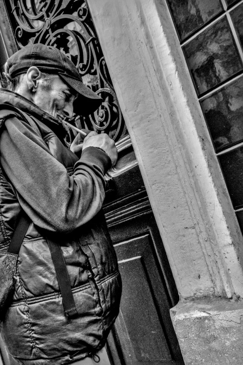 Frédéric · 11/04/2026 · 1120 x 1680 Cette photographie en noir et blanc met en scène un homme comme sujet principal, penché et absorbé par son téléphone portable. La composition est serrée, avec un cadrage rapproché qui isole le sujet. L’arrière-plan architectural, avec ses détails ornés et ses colonnes, ajoute une touche de profondeur et de contexte urbain.La lumière semble naturelle, douce et légèrement contrastée, créant des ombres marquées. Le style est documentaire et intime, capturant un moment de quotidien. L’ambiance est à la fois mélancolique et réaliste, renforcée par le monochrome qui accentue les textures et les jeux d’ombres.