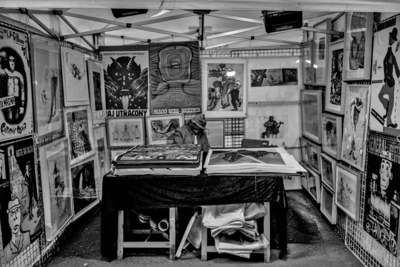 Frédéric · 11/04/2026 · 2517 x 1680 Cette photographie en noir et blanc montre une exposition d'art dans un espace confiné, probablement un train ou un wagon. Le sujet principal est une collection dense de tableaux et dessins accrochés aux murs et disposés sur des tables.La composition est serrée, avec un cadrage en plan moyen, mettant en valeur les œuvres et l'atmosphère intimiste. La lumière semble artificielle, créant des contrastes marqués. Les couleurs dominantes sont les nuances de gris, renforçant un style rétro ou vintage. L'ambiance est studieuse et artistique, évoquant un lieu dédié à la création et à l'exposition d'art.