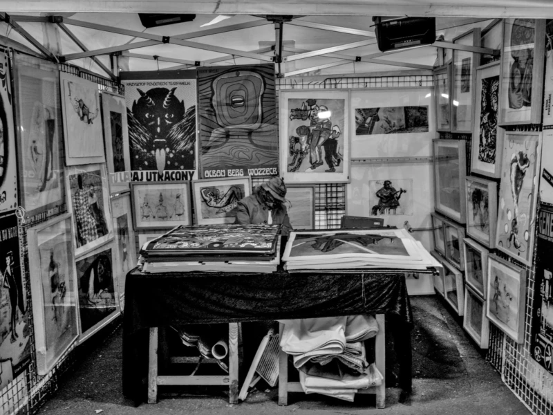 Frédéric · 11/04/2026 · 2517 x 1680 Cette photographie en noir et blanc montre une exposition d'art dans un espace confiné, probablement un train ou un wagon. Le sujet principal est une collection dense de tableaux et dessins accrochés aux murs et disposés sur des tables.La composition est serrée, avec un cadrage en plan moyen, mettant en valeur les œuvres et l'atmosphère intimiste. La lumière semble artificielle, créant des contrastes marqués. Les couleurs dominantes sont les nuances de gris, renforçant un style rétro ou vintage. L'ambiance est studieuse et artistique, évoquant un lieu dédié à la création et à l'exposition d'art.