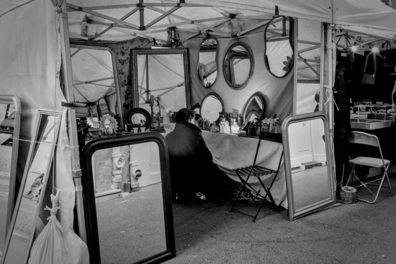 Frédéric · 11/04/2026 · 2517 x 1680 Cette photographie en noir et blanc montre un photographe en train de travailler dans un studio vintage. Le sujet principal est le photographe, assis derrière une grande glace réfléchissante.La composition met en scène plusieurs miroirs et accessoires rétro, créant une ambiance studieuse et artistique. Le cadrage est serré, centré sur le photographe et son environnement immédiat. La lumière semble douce et tamisée, typique des studios anciens. Les tons dominants sont les gris et noirs, renforçant l’aspect rétro et intemporel. Le style évoque une époque classique, avec une ambiance à la fois professionnelle et nostalgique.