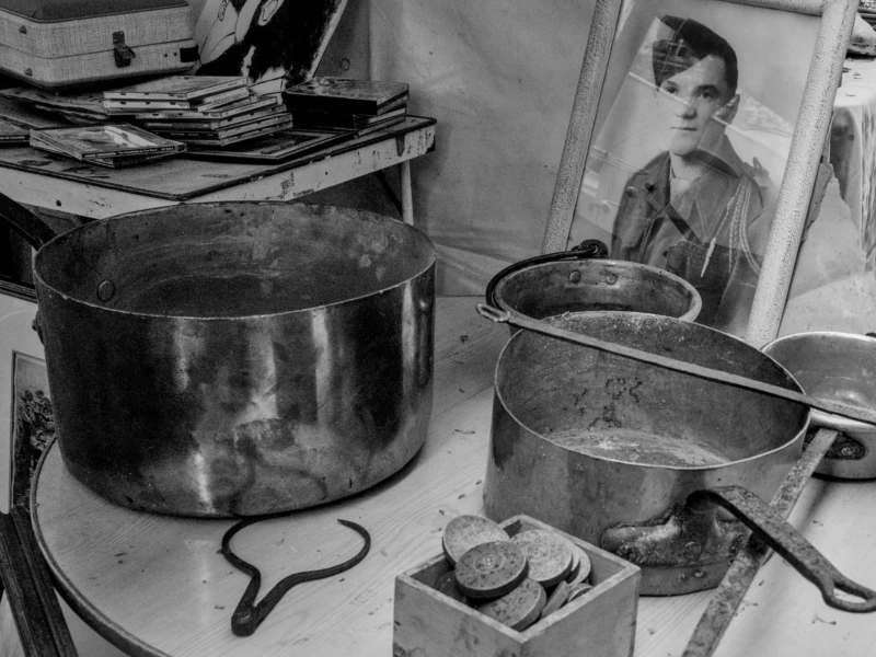 Frédéric · 11/04/2026 · 2517 x 1680 Cette photographie en noir et blanc montre une cuisine modeste. Le sujet principal est une femme reflétée dans un miroir, entourée de pots et casseroles en métal usés. La composition est divisée en deux parties : à gauche, les ustensiles de cuisine empilés, et à droite, la femme et son reflet.Le cadrage est serré, mettant en valeur les détails du quotidien. La lumière est douce et naturelle, créant des contrastes marqués. Les couleurs dominantes sont les nuances de gris, renforçant une ambiance sobre et réaliste. Le style est documentaire, évoquant une scène de vie authentique et intemporelle.