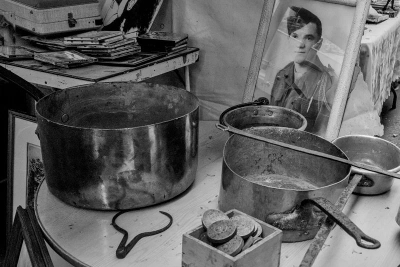 Frédéric · 11/04/2026 · 2517 x 1680 Cette photographie en noir et blanc montre une cuisine modeste. Le sujet principal est une femme reflétée dans un miroir, entourée de pots et casseroles en métal usés. La composition est divisée en deux parties : à gauche, les ustensiles de cuisine empilés, et à droite, la femme et son reflet.Le cadrage est serré, mettant en valeur les détails du quotidien. La lumière est douce et naturelle, créant des contrastes marqués. Les couleurs dominantes sont les nuances de gris, renforçant une ambiance sobre et réaliste. Le style est documentaire, évoquant une scène de vie authentique et intemporelle.
