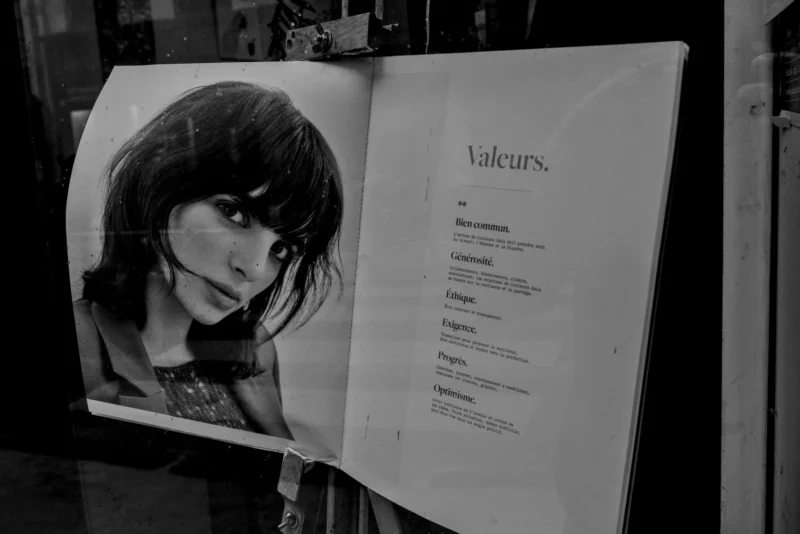 Frédéric · 11/04/2026 · 2517 x 1680 Cette photographie montre une affiche partiellement ouverte, avec pour sujet principal une jeune femme aux cheveux noirs, stylisée en noir et blanc. La composition met en valeur son portrait à gauche, tandis que la droite révèle une liste intitulée "Valeurs".Le cadrage est serré sur l'affiche, avec un angle légèrement oblique. La lumière est tamisée, créant des contrastes doux et des ombres marquées. Les couleurs dominantes sont les tons de gris, renforçant l'ambiance monochrome et mystérieuse. Le style est épuré et minimaliste, évoquant une esthétique moderne et introspective. L'ambiance est à la fois élégante et contemplative.