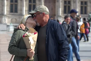 Fabienne · 3277 x 2188 Cette photographie capture un couple en train de s'embrasser tendrement. Le sujet principal est au centre, avec une femme tenant un bouquet de fleurs et un homme en veste sombre et chapeau. La composition est simple et naturelle, centrée sur l’émotion.Le cadrage est serré sur les deux personnages, avec un arrière-plan flou montrant des passants et un bâtiment classique. La lumière est douce et naturelle, probablement en extérieur. Les couleurs dominantes sont neutres (beige, gris) avec des touches de vert et de rouge dans les fleurs. L’ambiance est chaleureuse et romantique, évoquant une scène intime dans un cadre urbain.