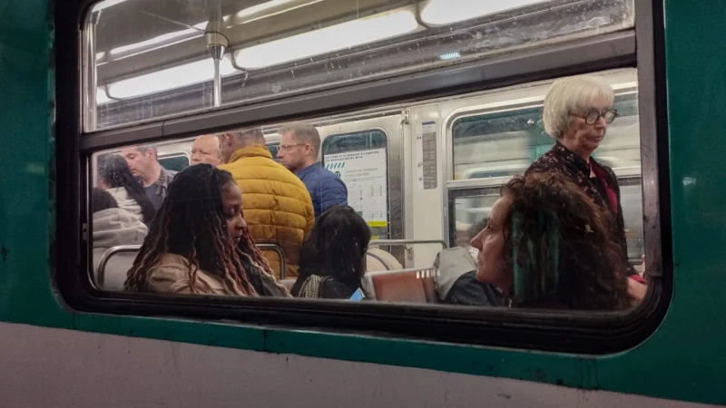 Muriel · 10/04/2026 · 2048 x 1152 Cette photographie capture l'intérieur d'un métro bondé. Le **sujet principal** est les passagers reflétés dans une vitre, créant une composition symétrique et dynamique.Le **cadrage** est serré, mettant en valeur les visages et les détails des voyageurs. La **lumière** est naturelle et tamisée, typique d’un intérieur éclairé artificiellement. Les **couleurs dominantes** sont neutres (gris, beige) avec des touches vives comme le jaune d’un vêtement.Le style est **documentaire et réaliste**, évoquant une scène du quotidien. L’**ambiance** est urbaine, animée et légèrement intimiste grâce au reflet.