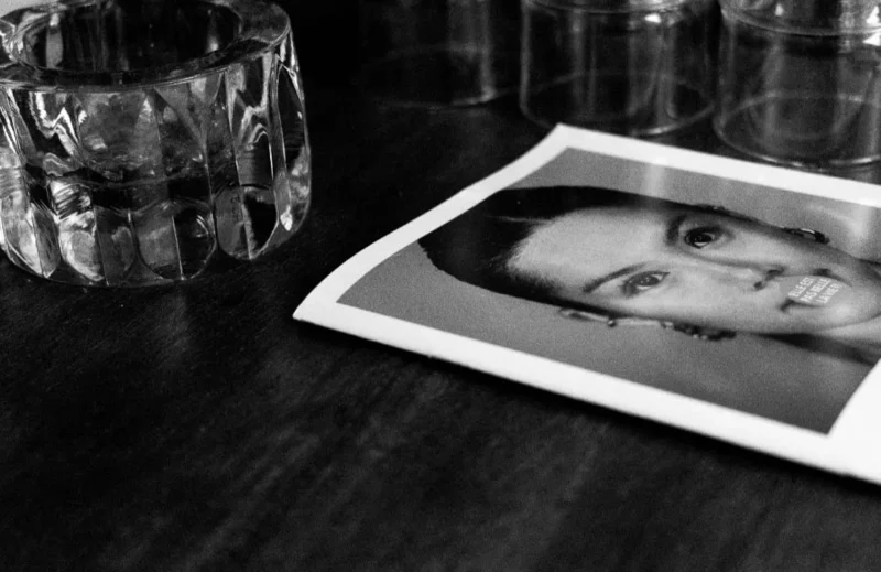 Muriel · 1080 x 700 Cette photographie en noir et blanc met en scène un portrait en pied d’une personne, partiellement visible, posé sur une surface sombre. Le sujet principal est ce cliché photographique, légèrement incliné.La composition est épurée, avec un cadrage serré sur le portrait et un arrière-plan minimaliste : une table et des verres en cristal. La lumière est douce, créant des contrastes subtils et des ombres discrètes. Les couleurs dominantes sont les nuances de gris, renforçant une ambiance intime et mélancolique. Le style est sobre et artistique, évoquant une touche nostalgique et contemplative.