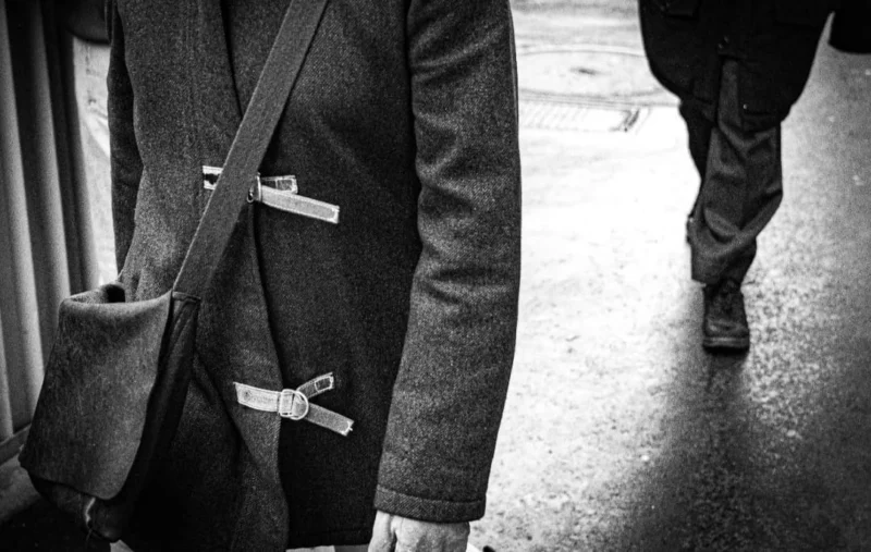 Muriel · 1080 x 685 Cette photographie en noir et blanc met en scène une personne de dos, vêtue d’un manteau avec des boutons en forme de nœuds papillon. Le sujet principal est flou, attirant l’attention sur les détails texturés du tissu et les boutons.La composition est simple et épurée, avec un cadrage serré sur le haut du corps. Les jambes d’une autre personne en mouvement sont partiellement visibles, ajoutant une touche de dynamisme. La lumière est douce, créant des contrastes subtils et des ombres discrètes. L’ambiance est intemporelle et élégante, renforcée par le style rétro et la sobriété des tons.
