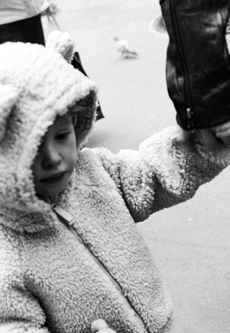 Muriel · 1080 x 1563 Cette photographie en noir et blanc montre une jeune enfant comme sujet principal, vêtue d’un épais manteau à capuche. Elle semble observer ou toucher quelque chose hors champ, avec un air concentré.La composition est simple et centrée sur son visage. Le cadrage est serré, mettant en valeur ses expressions et son regard. La lumière est douce et naturelle, sans contrastes marqués, créant une ambiance sobre et intime. Les tons dominants sont les gris et noirs, renforçant une atmosphère nostalgique et authentique. Le style est documentaire, évoquant une scène du quotidien. L’ambiance est à la fois douce et mélancolique.