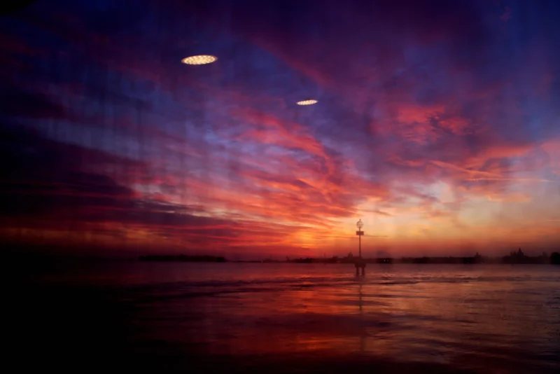 Fabienne · 09/04/2026 · 5488 x 3662 Cette photographie capture un coucher de soleil spectaculaire sur une étendue d’eau. Le sujet principal est le ciel aux teintes vibrantes, avec des nuages illuminés par des reflets orangés et violets. Deux formes lumineuses, probablement des avions ou des drones, traversent la scène.La composition est équilibrée, avec une ligne d’horizon centrale et des éléments verticaux (poteaux) qui guident le regard. Le cadrage large englobe l’immensité du paysage. Les couleurs dominantes sont les tons chauds (orange, rouge) contrastant avec les bleus et violets profonds.Le style est artistique et poétique, évoquant une ambiance paisible et presque mystique. La lumière douce et diffuse accentue l’émotion.