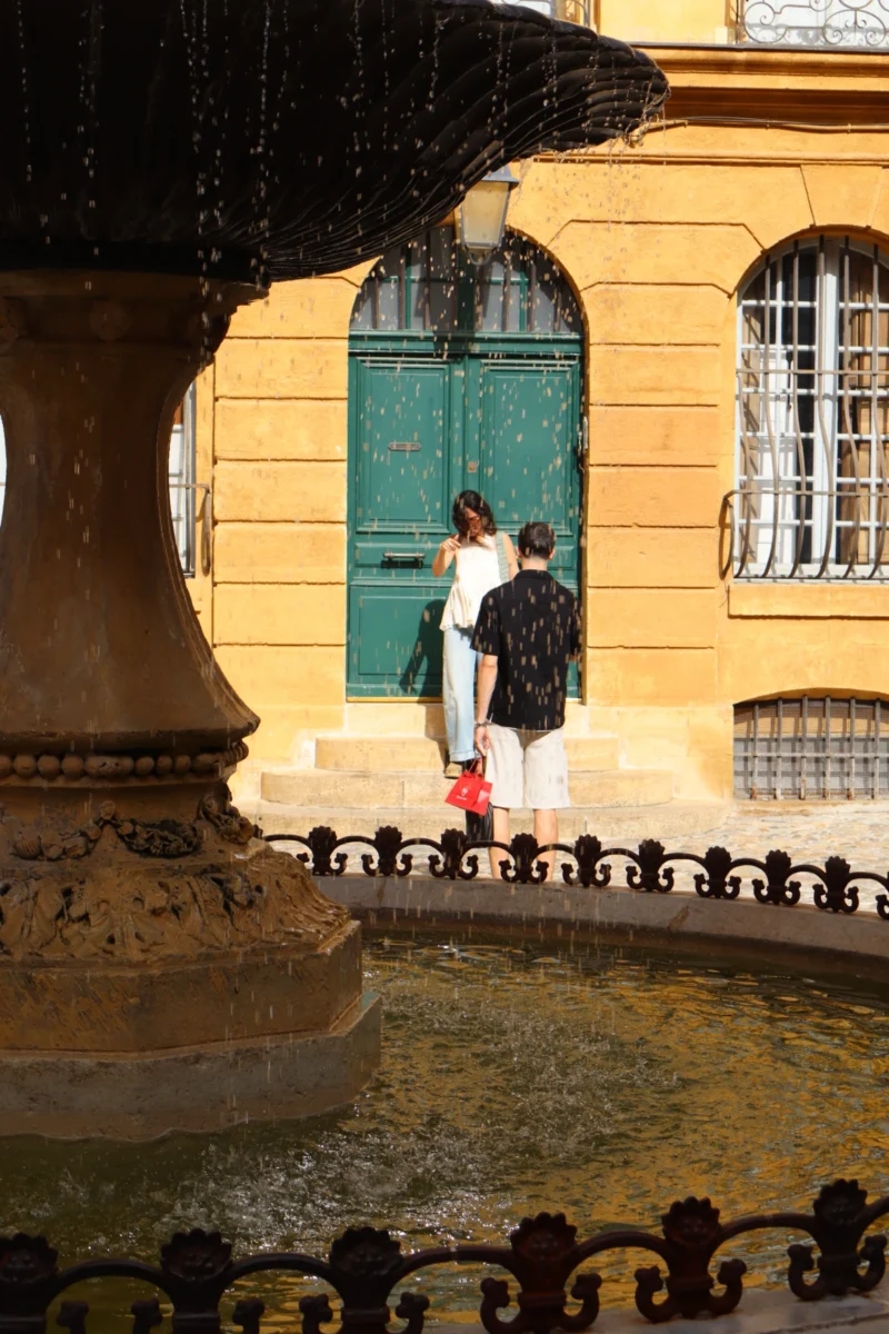 Camilla · 3971 x 5956 Cette photographie met en scène deux enfants comme sujets principaux, debout près d’une fontaine en pierre. La composition est équilibrée, avec la fontaine en premier plan et un bâtiment jaune en arrière-plan. Le cadrage est serré sur les enfants, isolant leur silhouette.La lumière semble naturelle, douce et légèrement tamisée, créant des ombres douces. Les couleurs dominantes sont les tons chauds (jaune du bâtiment) et les teintes froides (vert des fenêtres, gris de la pierre). Le style est réaliste et documentaire, évoquant une ambiance paisible et presque nostalgique, typique d’un moment de jeu ou d’exploration urbaine.
