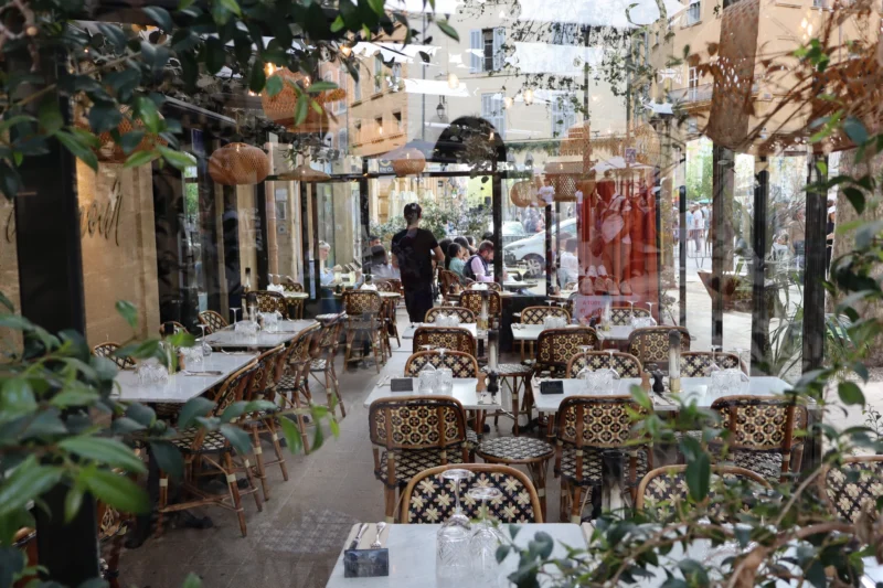 Camilla · 5955 x 3970 Cette photographie montre une terrasse de restaurant extérieure. Le sujet principal est l’espace aménagé avec des tables et chaises en rotin, couvertes de nappes blanches. La composition est symétrique, avec des plantes vertes et des éléments décoratifs en arrière-plan.Le cadrage est large, incluant une allée centrale et des détails architecturaux. La lumière semble naturelle, douce, créant des ombres légères. Les couleurs dominantes sont les verts des plantes, les tons neutres des tables et les touches de terre cuite des murs.L’ambiance est calme et élégante, évoquant un lieu convivial et raffiné.