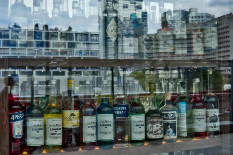 Frédéric · 19/04/2026 · 2516 x 1680 Cette photographie montre une collection de bouteilles de whisky alignées sur un rebord, avec des bougies allumées devant chacune. Le sujet principal est ces bouteilles, variées en couleurs et en marques.La composition est horizontale, avec un cadrage serré sur les bouteilles en premier plan, tandis qu’en arrière-plan, des bâtiments urbains flous forment un fond esthétique. La lumière est douce et naturelle, probablement en journée, créant des ombres légères. Les couleurs dominantes sont les tons bruns des bouteilles, contrastant avec les teintes claires des bougies et des bâtiments.L’ambiance est à la fois chaleureuse et nostalgique, évoquant une célébration ou un hommage.