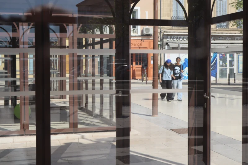 Camilla · 26/04/2026 · 6000 x 4000 Cette photographie montre un espace urbain vu à travers une structure vitrée et métallique. Le sujet principal semble être une allée couverte avec des colonnes et des bâtiments en arrière-plan.La composition repose sur des jeux de reflets et de transparences, créant une ambiance presque labyrinthique. Le cadrage est serré, centré sur les éléments architecturaux et les reflets.La lumière est naturelle, probablement en journée, avec des ombres douces. Les couleurs dominantes sont des tons neutres (gris, beige) et quelques touches de bleu et vert.Le style est contemporain et minimaliste, évoquant une ambiance à la fois moderne et légèrement mystérieuse.