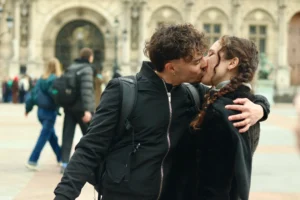 Francoise · 04/04/2026 · 6000 x 4000 Cette photographie met en scène un couple qui s'embrasse tendrement en extérieur. Le sujet principal est centré sur eux, avec un cadrage serré qui isole leur moment intime. L'arrière-plan flou montre des passants et une architecture classique, suggérant une place publique.La lumière est douce et naturelle, probablement en journée. Les couleurs dominantes sont neutres et froides, avec des tons gris et noirs, adoucis par les vêtements sombres du couple. L'ambiance est romantique et intimiste malgré le cadre urbain. Le style est réaliste et spontané.