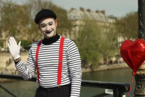 Francoise · 04/04/2026 · 6000 x 4000 Cette photographie met en scène un mime en costume traditionnel : haut rayé noir et blanc, pantalon blanc, souliers rouges et chapeau noir. Le sujet principal est centré, avec un cadrage serré qui isole son visage et ses gestes expressifs (sourire et geste de la main).La lumière naturelle éclaire le sujet de face, créant des contrastes doux. Les couleurs dominantes sont les tons blancs, noirs et rouges, renforçant le style clownesque. L’arrière-plan flou, avec des bâtiments et des arbres, ajoute une ambiance urbaine et légère, tout en mettant en valeur le personnage. L’ensemble dégage une touche à la fois joyeuse et nostalgique.