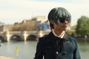 Francoise · 04/04/2026 · 6000 x 4000 Cette photographie met en scène une personne au style androgyne et mystérieux, vêtue d’un manteau bleu marine avec des boutons dorés, une cravate et des cheveux noirs striés. Le cadrage est serré sur son visage partiellement masqué par des lunettes et une mèche tombante.L’arrière-plan montre un pont emblématique et une rivière, suggérant un cadre urbain. La lumière est douce, naturelle, avec des ombres légères, créant une ambiance à la fois sobre et dramatique. Les couleurs dominantes sont les bleus, gris et noirs, renforçant un style sombre et élégant, évoquant une touche cinématographique ou gothique.