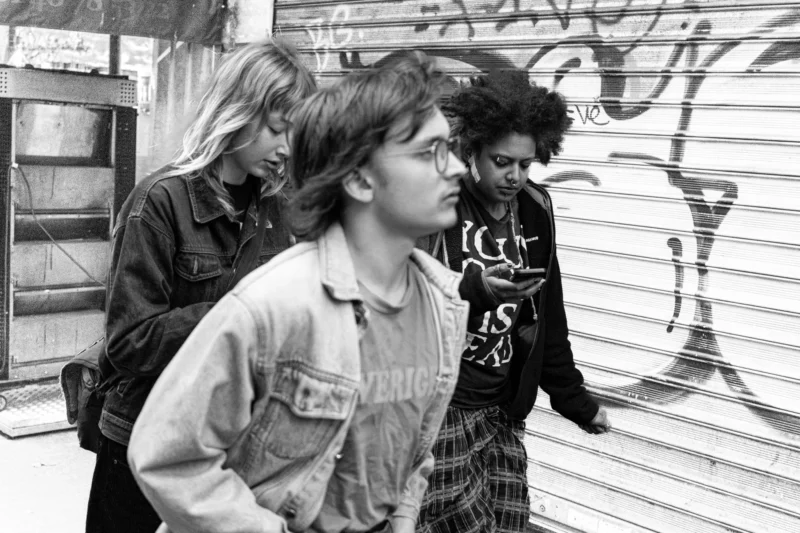 Muriel · 11/04/2026 · 2048 x 1365 Cette photographie en noir et blanc montre trois jeunes hommes marchant côte à côte. Le sujet principal semble être le personnage central, vêtu d'une veste en jean et d'un pull à capuche.La composition est dynamique, avec un cadrage serré sur les visages et les épaules. La lumière est naturelle et semble provenir de la gauche, créant des contrastes doux. L’ambiance est urbaine et informelle, évoquant une époque rétro, probablement les années 1980 ou 1990. Le style est documentaire, capturant un moment authentique et spontané.