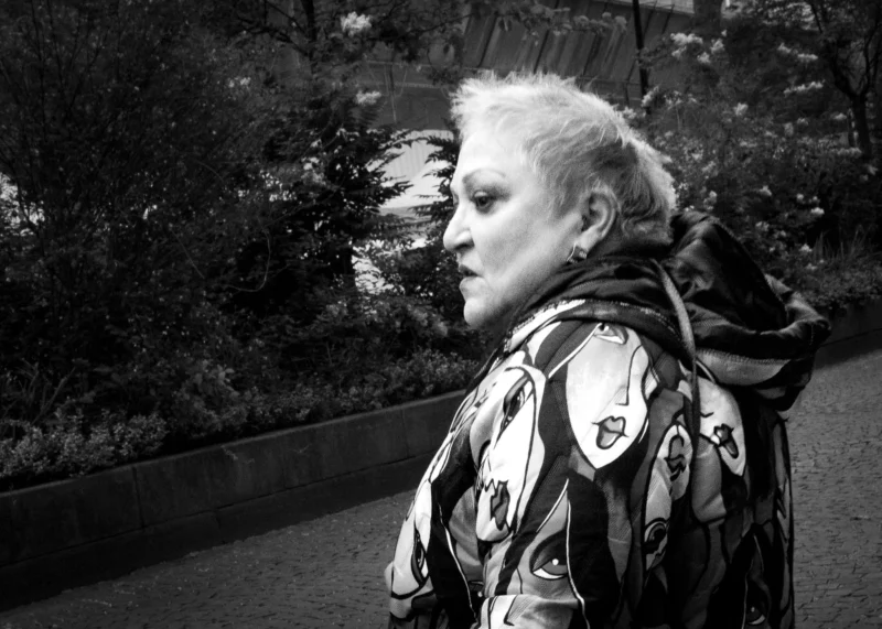 Muriel · 11/04/2026 · 2048 x 1463 Cette photographie met en scène une femme âgée comme sujet principal, vue de profil. La composition joue sur le flou d'arrière-plan, mettant en valeur son visage et ses traits expressifs. Le cadrage serré et légèrement bas accentue son regard et son expression mélancolique.La lumière est douce, créant des contrastes subtils. En noir et blanc, les tons varient du clair au foncé, renforçant les textures du vêtement et des cheveux.Le style est documentaire et intime, évoquant une ambiance contemplative et presque poétique.