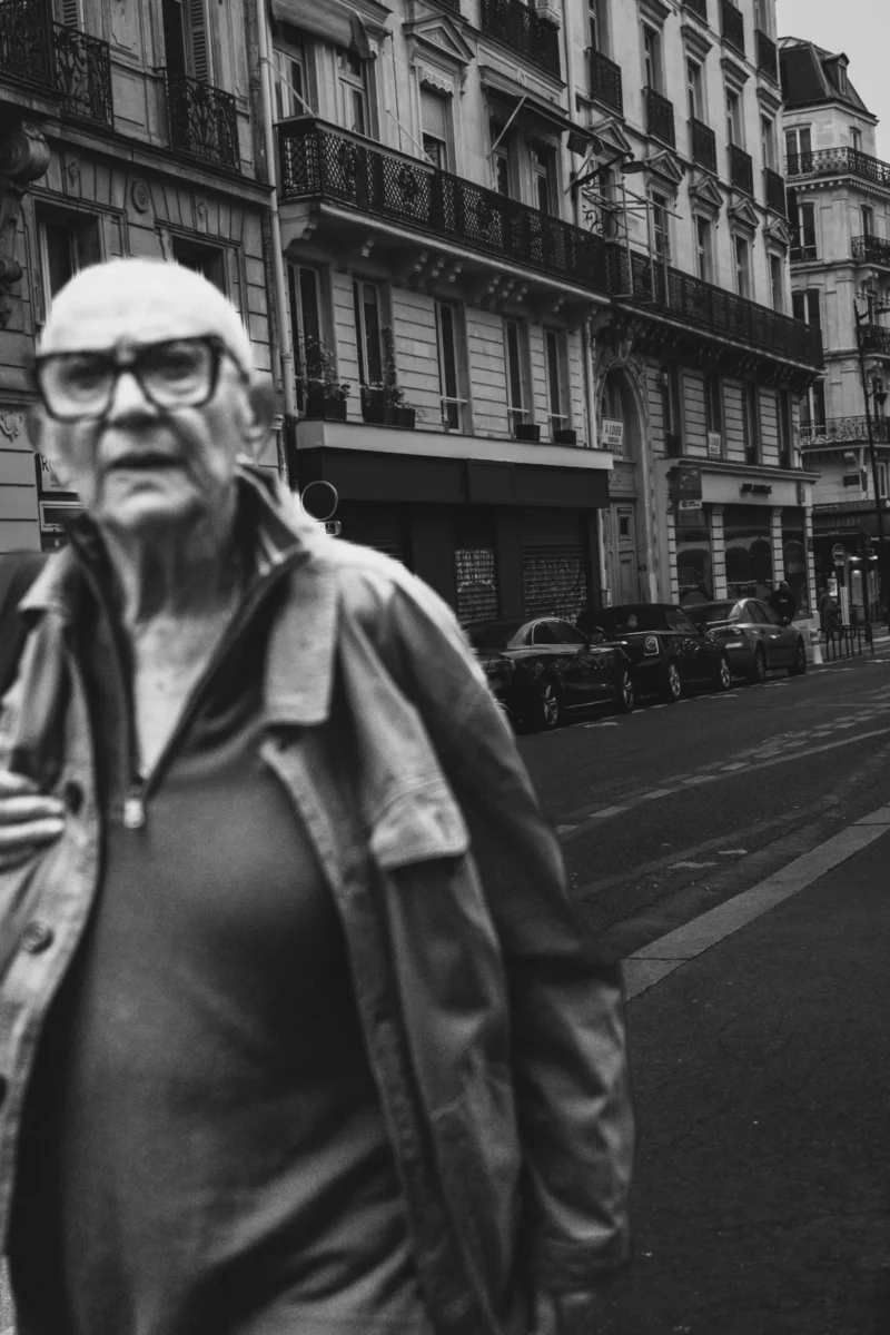 Muriel · 11/04/2026 · 3700 x 5550 Cette photographie en noir et blanc met en scène un vieil homme comme sujet principal, vêtu d’un manteau et d’un chapeau, marchant sur un trottoir urbain. La composition est simple et naturelle, avec un cadrage serré sur le sujet, laissant en arrière-plan des bâtiments anciens et une rue étroite.La lumière, douce et légèrement contrastée, crée des ombres marquées, accentuant les textures. Les tons dominants sont les gris et noirs, typiques du noir et blanc, renforçant une ambiance mélancolique et intemporelle. Le style est documentaire et sobre, évoquant une atmosphère à la fois calme et nostalgique.