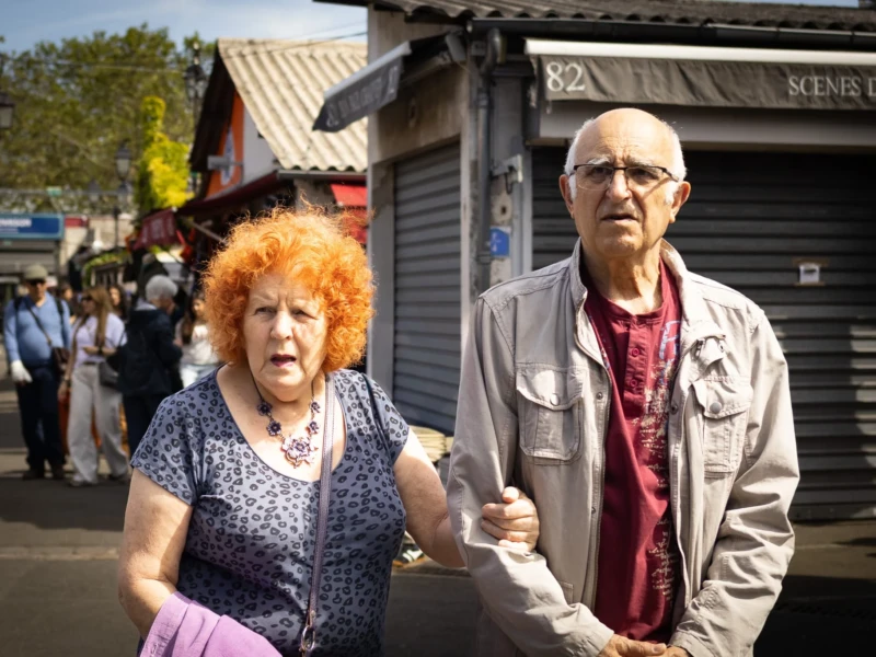 Muriel · 25/04/2026 · 2048 x 1365 Cette photographie met en scène un couple comme sujets principaux, marchant côte à côte. La femme, avec ses cheveux roux et un pull à motifs, et l’homme, vêtu d’une veste beige, semblent engagés dans une conversation.La composition est simple et naturelle, avec un cadrage serré sur eux. L’arrière-plan flou montre une rue animée, avec des bâtiments et des passants, créant une ambiance urbaine.La lumière est naturelle et semble provenir d’un jour ensoleillé, apportant des contrastes doux. Les couleurs dominantes sont les tons neutres et naturels, avec des touches de rouge et de bleu. Le style est documentaire et réaliste, évoquant une ambiance quotidienne et authentique.