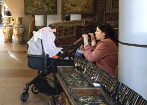 Muriel · 25/04/2026 · 2048 x 1463 Cette photographie montre une femme assise dans un lieu qui semble être un musée ou une boutique d'artisanat, tenant un téléphone à l'oreille. Le sujet principal est cette femme, entourée de divers objets artisanaux et textiles colorés.La composition met en valeur un cadrage serré sur elle, avec un fond riche en motifs et couleurs. La lumière est naturelle et douce, éclairant les détails des objets et créant des ombres légères. Les couleurs dominantes sont chaudes et variées, avec des teintes terreuses et des touches de rouge et bleu.L’ambiance est calme et intime, évoquant une pause contemplative dans un environnement culturel et artisanal.