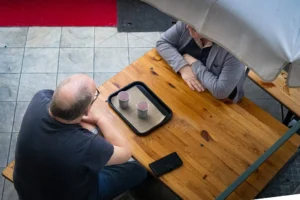 Muriel · 25/04/2026 · 2048 x 1365 Cette photographie montre deux hommes assis à une table en bois, discutant. Le sujet principal est leur interaction, avec un cadrage en plan serré sur la table et leurs mains.La composition est symétrique et centrée, mettant en valeur l’espace entre eux. La lumière est naturelle et douce, éclairant uniformément la scène. Les couleurs dominantes sont les tons neutres (bois, gris) avec des touches de rouge et blanc en arrière-plan.Le style est sobre et réaliste, évoquant une ambiance calme et professionnelle, presque intime.