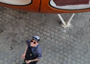 Muriel · 25/04/2026 · 2048 x 1463 Cette photographie montre un homme en position centrale, vu d’en haut, comme sujet principal. Il porte une veste bleue, un chapeau et des lunettes de soleil, donnant un air décontracté.La composition est symétrique avec un cadrage serré, mettant en valeur le sujet sous un abri bus orange. La lumière semble naturelle, bien que légèrement contrastée, créant des ombres douces. Les couleurs dominantes sont les tons gris du sol pavé, le bleu de la veste et l’orange vif de l’abri.Le style est documentaire et spontané, évoquant une ambiance urbaine et quotidienne, presque intimiste malgré l’absence d’expression visible du visage.