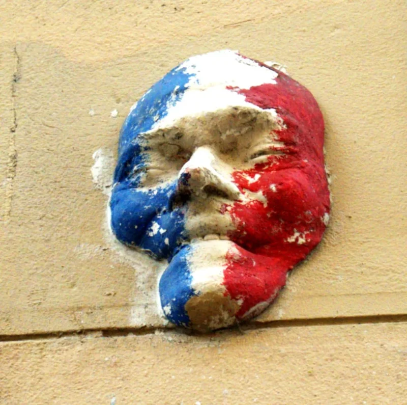 Bao · 1131 x 1124 Cette photographie montre un visage stylisé peint sur un mur, évoquant une tête déformée aux couleurs vives. Les tons dominants sont le bleu, blanc et rouge, rappelant les couleurs du drapeau français.Le cadrage est serré, centré sur le sujet. La lumière semble naturelle, mettant en valeur les textures et les dégradés de peinture. Le style est brut et expressif, typique du street art. L’ambiance est à la fois mystérieuse et provocante, avec une touche d’humour noir.