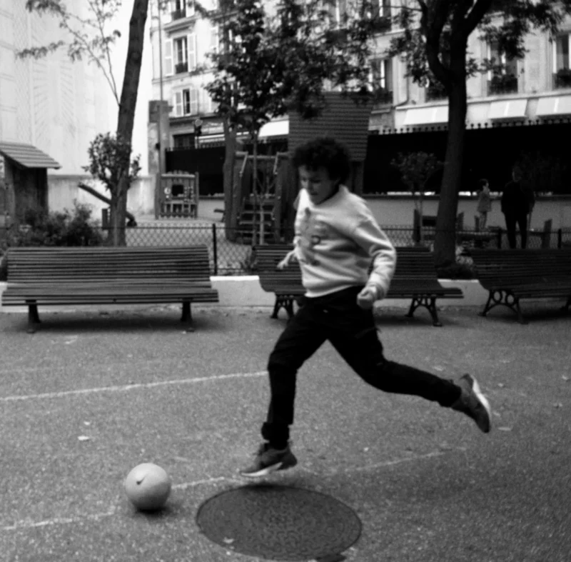 Bao · 3493 x 3438 Cette photographie en noir et blanc capture un jeune homme en mouvement, sujet principal, jouant au football dans un espace urbain. La composition met en valeur son action dynamique, avec un cadrage serré qui isole le sujet du premier plan.La lumière est naturelle, probablement en journée, créant des contrastes nets. L’ambiance est à la fois spontanée et nostalgique, renforcée par le style rétro du noir et blanc. Les couleurs dominantes, bien que monochromes, suggèrent des tons gris et blancs, avec des ombres marquées. L’arrière-plan, flou et discret, accentue l’énergie du sujet.