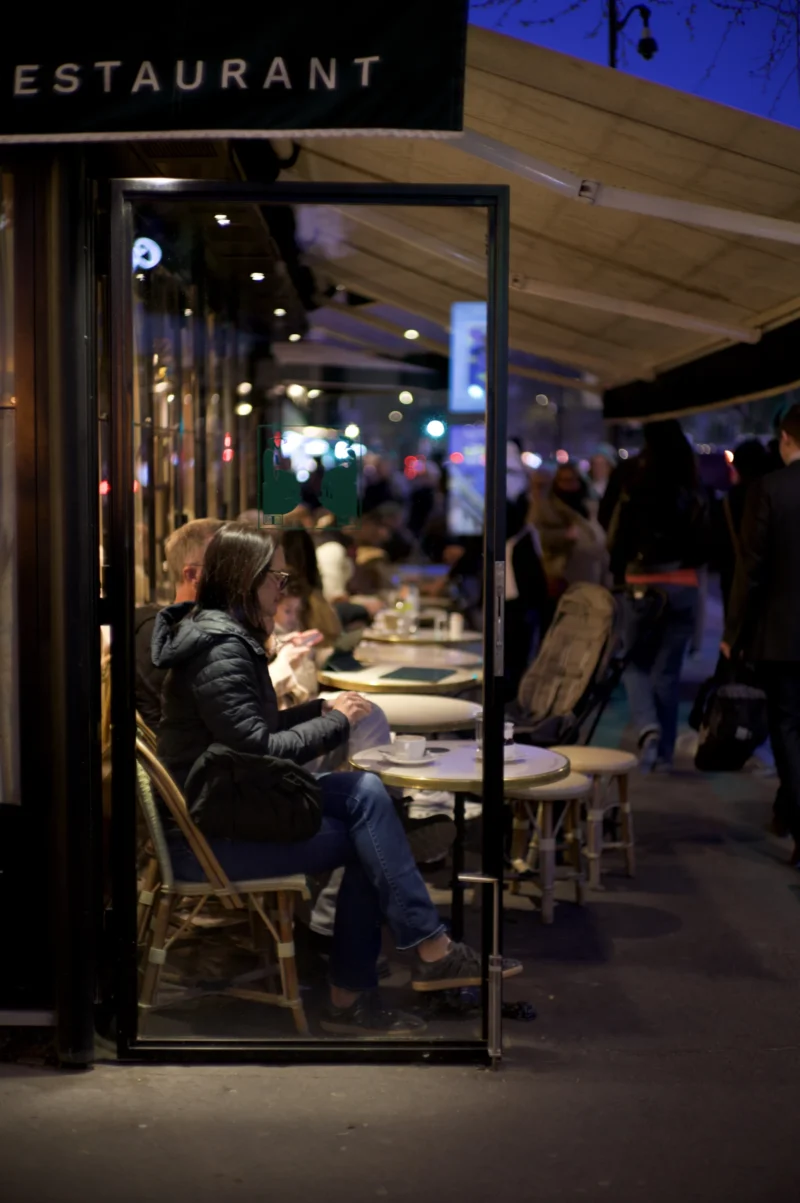 Thomas · 30/03/2026 · 2724 x 4096 Cette photographie montre un **restaurant extérieur** la nuit. Le **sujet principal** est un couple assis à une table, éclairés par une lumière douce. La **composition** met en valeur les tables et les clients en arrière-plan, avec un cadrage serré sur le couple en premier plan.La **lumière** est tamisée, créant une ambiance chaleureuse et intimiste. Les **couleurs dominantes** sont des tons sombres (noir, gris) rehaussés par des éclairages jaunâtres et blancs.Le **style** est réaliste et documentaire, capturant une scène de vie urbaine. L’**ambiance** est conviviale et légèrement nocturne, évoquant une soirée détendue en ville.