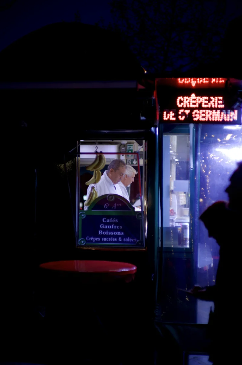 Thomas · 30/03/2026 · 2724 x 4096 Cette photographie montre un stand de crêpes éclairé la nuit, intitulé "Crêperie du Cœur Sauvage". Le sujet principal est une personne derrière le comptoir, en train de préparer des crêpes.La composition est centrée sur le stand, avec un cadrage serré qui met en valeur les détails lumineux. La lumière est douce et artificielle, créant une ambiance chaleureuse et conviviale. Les couleurs dominantes sont des tons bleus nocturnes, contrastant avec les éclairages chauds jaunes et rouges du stand.Le style est réaliste et spontané, évoquant une scène nocturne urbaine et authentique.