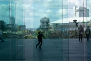 Laurence · 28/03/2026 · 1350 x 900 Cette photographie montre deux personnes reflétées dans une surface vitrée transparente. Le sujet principal semble être le contraste entre leur mouvement et leur reflet.La composition utilise une perspective symétrique et horizontale, avec un cadrage serré sur le sol vitré. La lumière est naturelle, douce et légèrement diffuse, créant des ombres subtiles. Les couleurs dominantes sont des teintes froides (bleutées et grises) avec des touches de vert et de blanc.Le style est épuré et moderne, évoquant une ambiance urbaine contemporaine et presque onirique, mêlant réalité et réflexion.