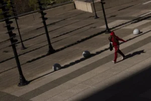 Laurence · 28/03/2026 · 1350 x 900 Cette photographie capture une personne en mouvement, vêtue d’une combinaison rouge, traversant une place urbaine. Le sujet principal est centré, avec une composition axée sur les lignes droites des lampadaires et des pavés.Le cadrage est serré, mettant en valeur le contraste entre la silhouette et l’architecture. La lumière, naturelle et rasante, crée des ombres marquées, accentuant la profondeur. Les couleurs dominantes sont les tons gris des pavés et le rouge vif de la combinaison, apportant une touche dynamique.Le style est épuré et documentaire, évoquant une ambiance à la fois urbaine et intemporelle, presque cinématographique.