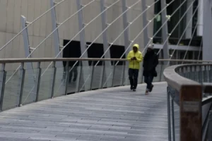Laurence · 28/03/2026 · 1350 x 900 Cette photographie montre deux personnes marchant sur un pont moderne. Le sujet principal est l’une d’elles, vêtue d’un manteau jaune fluo, qui attire l’attention par sa couleur vive.La composition repose sur une perspective dynamique, avec des lignes métalliques et des garde-corps en diagonale. Le cadrage est serré, mettant en valeur les sujets et l’architecture environnante. La lumière semble naturelle mais diffuse, créant une ambiance sobre et légèrement grise. Les couleurs dominantes sont les tons froids (gris, bleu) contrastés par le jaune vif du manteau.Le style est épuré et contemporain, évoquant une ambiance urbaine et moderne.