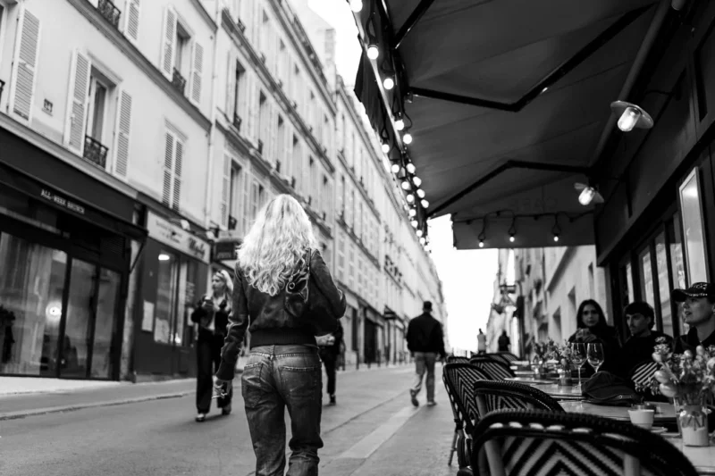 Laurence · 11/04/2026 · 1350 x 900 Cette photographie en noir et blanc met en scène une femme au premier plan, vêtue d’une robe ample et d’un chapeau, marchant dans une rue urbaine. La composition joue sur la profondeur avec des bâtiments alignés et des passants en arrière-plan.Le cadrage est serré sur la femme, mais inclut aussi des éléments architecturaux et des détails de la vie quotidienne. La lumière, douce et naturelle, crée des contrastes subtils. Les couleurs dominantes, bien que monochromes, varient en intensité grâce aux jeux d’ombres et de lumières.Le style est documentaire et réaliste, évoquant une ambiance calme et mélancolique, typique d’une scène de rue parisienne.