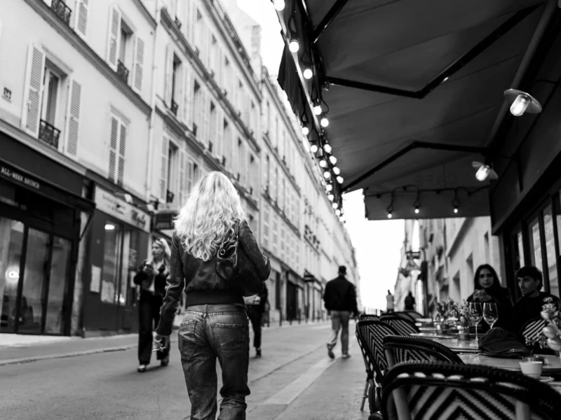Laurence · 11/04/2026 · 1350 x 900 Cette photographie en noir et blanc met en scène une femme au premier plan, vêtue d’une robe ample et d’un chapeau, marchant dans une rue urbaine. La composition joue sur la profondeur avec des bâtiments alignés et des passants en arrière-plan.Le cadrage est serré sur la femme, mais inclut aussi des éléments architecturaux et des détails de la vie quotidienne. La lumière, douce et naturelle, crée des contrastes subtils. Les couleurs dominantes, bien que monochromes, varient en intensité grâce aux jeux d’ombres et de lumières.Le style est documentaire et réaliste, évoquant une ambiance calme et mélancolique, typique d’une scène de rue parisienne.
