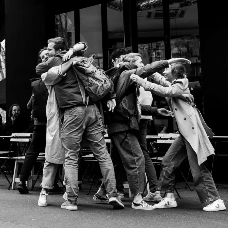 Laurence · 11/04/2026 · 900 x 900 Cette photographie capture un groupe de personnes en mouvement, probablement en train de danser ou de célébrer. Le sujet principal est l’énergie collective et la spontanéité du groupe.La composition est dynamique, avec un cadrage serré sur les individus en interaction. La lumière est naturelle, contrastée, créant des ombres marquées. En noir et blanc, les tons dominants varient du clair au foncé, accentuant les textures des vêtements et les expressions.Le style est documentaire et spontané, évoquant une ambiance joyeuse et vivante, presque festive.
