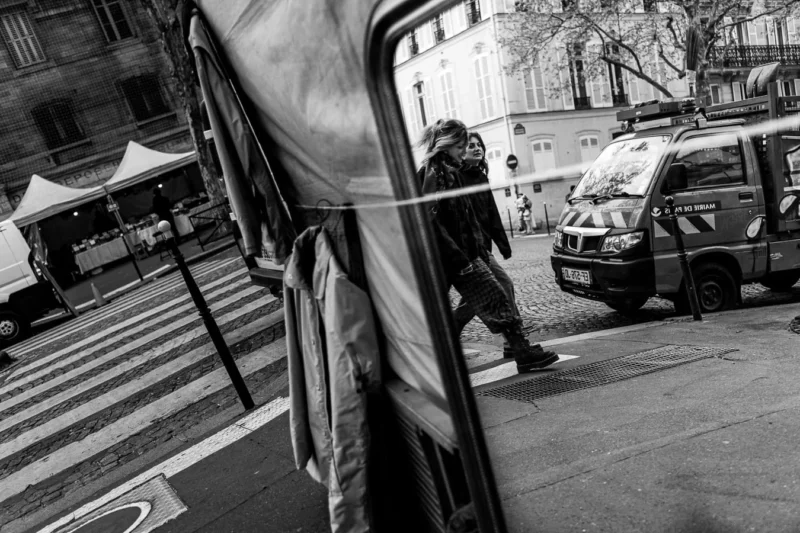 Laurence · 11/04/2026 · 1350 x 900 Cette photographie en noir et blanc met en scène un homme marchant avec une grande bâche transparente. Le sujet principal est flou, suggérant un mouvement rapide.La composition joue sur les contrastes entre les formes géométriques (trottoir, bâche) et les éléments urbains (voitures, bâtiments). Le cadrage serré et légèrement bas accentue la dynamique et l’anonymat du personnage.La lumière, douce et diffuse, crée des ombres marquées, renforçant l’ambiance urbaine et mélancolique. Les tons dominants, du noir au gris, soulignent les textures et les jeux d’ombres. Le style documentaire confère une touche réaliste et intemporelle.