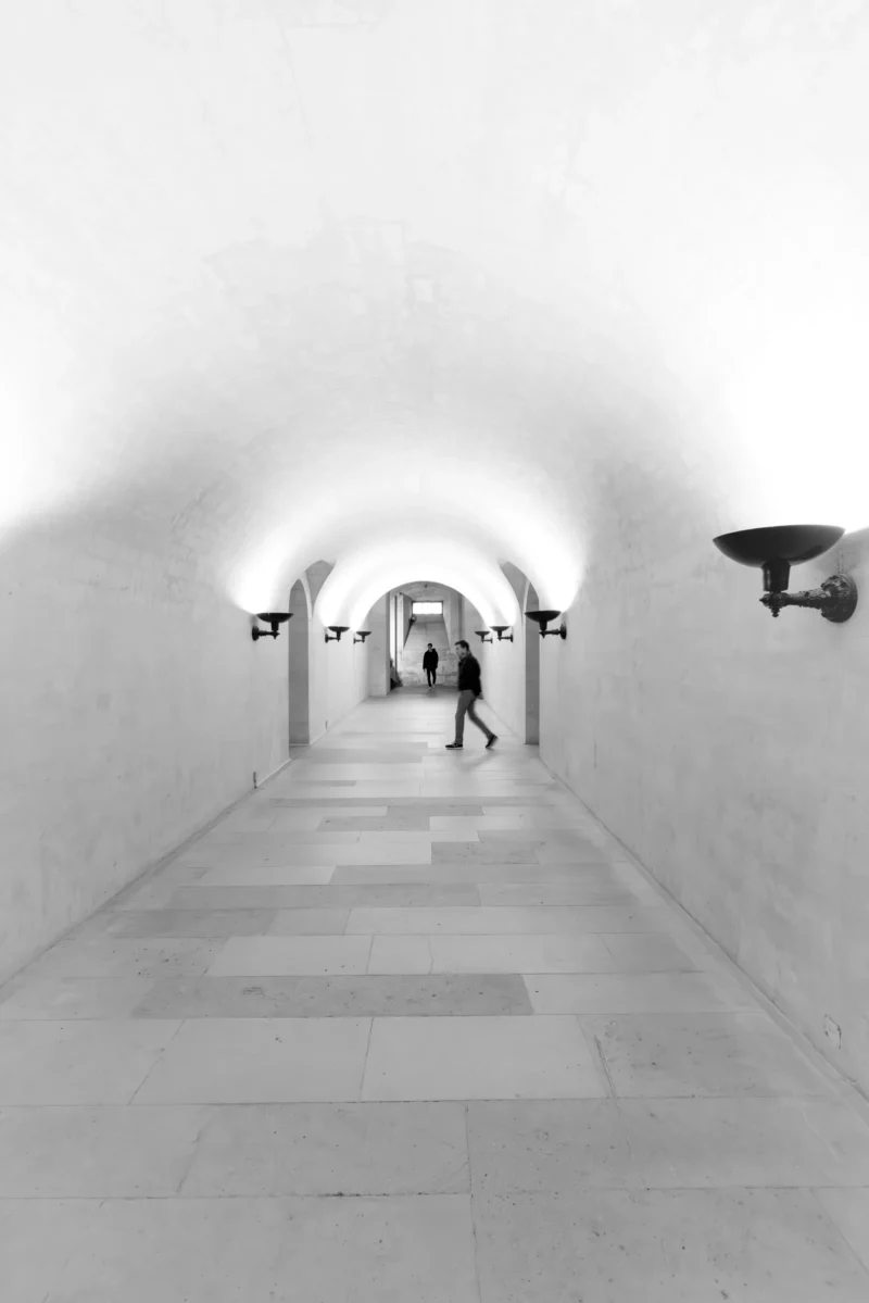 Philippe · 2001 x 3000 Cette photographie en noir et blanc montre un couloir souterrain ou industriel. Le sujet principal est une personne en mouvement, au centre, éclairée par une lumière douce en provenance de l’extrémité opposée.La composition est symétrique, avec des murs blancs et un sol carrelé. Le cadrage est serré, guidant le regard vers le fond. La lumière, tamisée et contrastée, crée une ambiance mystérieuse et minimaliste. Les tons dominants sont les gris et blancs, renforçant une atmosphère sobre et épurée. Le style est documentaire, évoquant une sensation de solitude et de profondeur.