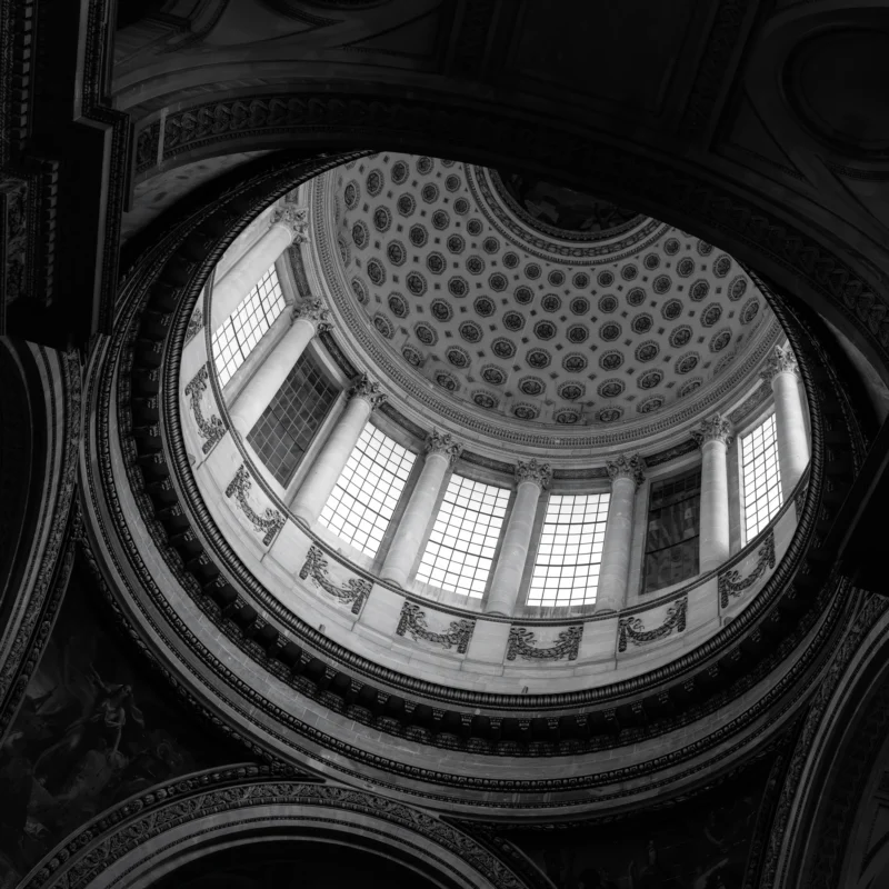 Philippe · 3000 x 3000 Cette photographie met en scène une **coupole architecturale** imposante, probablement d’un bâtiment historique. Le **sujet principal** est la structure géométrique et les **fenêtres en forme de losange** laissant filtrer la lumière.La **composition** est centrée, avec un **cadrage serré** sur la voûte, accentuant sa grandeur. La **lumière**, naturelle et contrastée, crée des ombres marquées, soulignant les détails. En **noir et blanc**, les tons varient du clair au très foncé, renforçant le **style dramatique et classique**.L’**ambiance** est solennelle, presque mystique, évoquant l’élégance et la permanence des grands édifices.