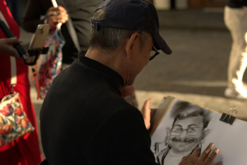 Bao · 6024 x 4016 Cette photographie montre un homme, sujet principal, penché sur une feuille où il dessine ou signe un portrait caricaturé. Le cadrage est serré sur son visage et ses mains, avec un arrière-plan flou.La lumière semble naturelle, douce et légèrement tamisée. Les couleurs dominantes sont sobres : noir (vêtements, chapeau), gris (dessins), avec des touches de rouge et de motifs colorés en arrière-plan.L’ambiance est intimiste et artistique, évoquant un marché ou une foire où l’artiste travaille. Le style est réaliste et spontané, reflétant un moment de création.