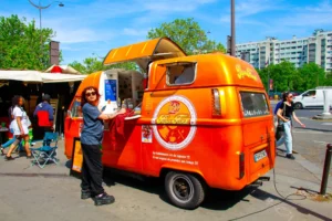 Cédric · 25/04/2026 · 2048 x 1365 Cette photographie met en scène un **food truck orange vif** comme sujet principal, avec un logo distinctif. La composition montre des personnes en mouvement autour du véhicule, dans une ambiance animée.Le cadrage est serré sur le camion, avec des éléments urbains en arrière-plan. La lumière est naturelle et éclatante, suggérant un jour ensoleillé. Les **couleurs dominantes** sont l’orange du camion, contrastant avec les tons neutres de l’environnement.Le style est dynamique et informel, évoquant une scène de rue vivante, typique d’un marché ou d’une fête populaire. L’ambiance est conviviale et énergique.