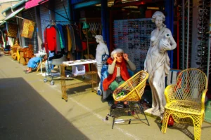 Cédric · 25/04/2026 · 2048 x 1365 Cette photographie montre un marché artisanal coloré. Le sujet principal semble être une femme assise sur un tabouret en train de travailler, entourée d’objets artisanaux.La composition est dynamique, avec des étals remplis de tissus et accessoires colorés en arrière-plan. Le cadrage est serré sur la scène centrale, mais inclut aussi des éléments périphériques.La lumière est vive et naturelle, mettant en valeur les couleurs vives. Les teintes dominantes sont les tons chauds (rouges, jaunes, oranges) et les contrastes entre les objets artisanaux et les statues.L’ambiance est vivante et artisanale, évoquant un marché traditionnel et chaleureux.