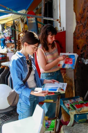 Cédric · 25/04/2026 · 1365 x 2048 Cette photographie montre deux jeunes femmes examinant des livres ou des magazines dans un marché ou une brocante. Le sujet principal est leur interaction avec les documents.La composition est dynamique, avec un cadrage serré sur les deux femmes. La lumière naturelle éclaire la scène, mettant en valeur les détails. Les couleurs dominantes sont vives et variées : bleu, rouge, vert et tons pastel.Le style est documentaire et spontané, capturant un moment authentique. L’ambiance est conviviale et décontractée, évoquant une recherche ou une découverte commune.